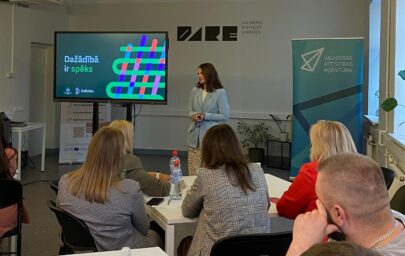 Sanita Lāce, Sabiedrības integrācijas fonda Dažādības vadības un ilgtspējas nodaļas vadītāja, vada prezentāciju Valmieras koprades darbnīcā. Uz ekrāna redzams uzraksts “Dažādībā ir spēks” ar krāsainu dizainu. Auditorija sēž pie galdiem, klausās un iesaistās diskusijā. Telpā redzams arī “Valmieras attīstības aģentūras” baneris.