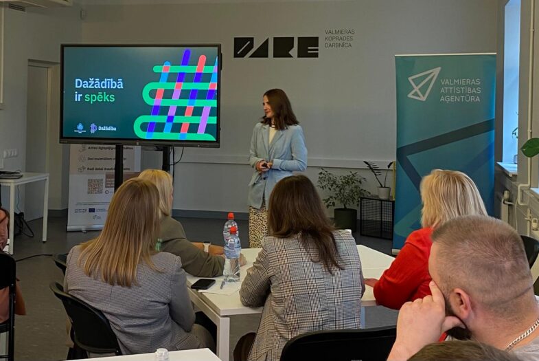 Sanita Lāce, Sabiedrības integrācijas fonda Dažādības vadības un ilgtspējas nodaļas vadītāja, vada prezentāciju Valmieras koprades darbnīcā. Uz ekrāna redzams uzraksts “Dažādībā ir spēks” ar krāsainu dizainu. Auditorija sēž pie galdiem, klausās un iesaistās diskusijā. Telpā redzams arī “Valmieras attīstības aģentūras” baneris.