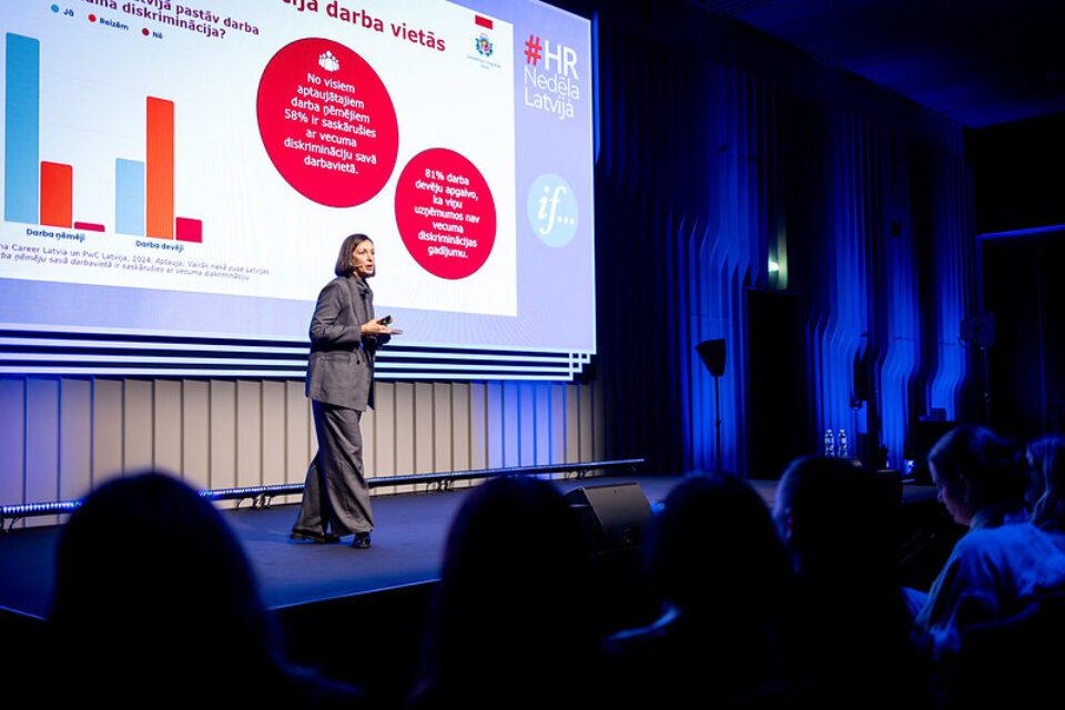 SIF sekretariāta direktore Inese Kalvāne sniedz prezentāciju uz skatuves. Aptumšotā zālē sēž skatītāji