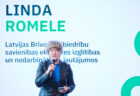 Sieviete ar mikrofonu uzstājas pasākumā, fonā projicēts viņas vārds “Linda Romele” un informācija par pārstāvētajām jomām.