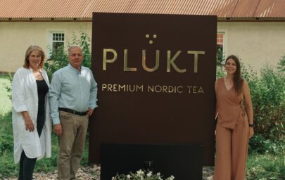 Trīs cilvēki stāv pie brūna “PLŪKT Premium Nordic Tea” zīmola stenda lauku vidē pie ēkas ar dzeltenu fasādi un brūnu jumtu.