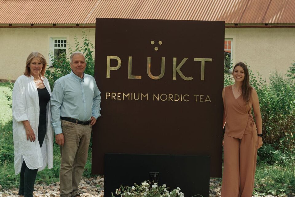Trīs cilvēki stāv pie brūna “PLŪKT Premium Nordic Tea” zīmola stenda lauku vidē pie ēkas ar dzeltenu fasādi un brūnu jumtu.