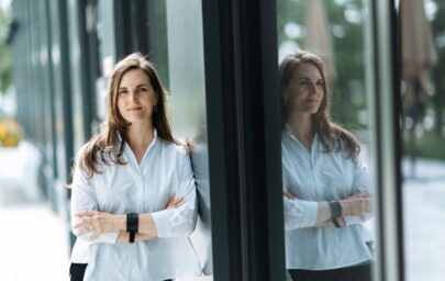 Sieviete baltā kreklā stāv pie mūsdienīgas stikla ēkas sienas ar sakrustotām rokām; stiklā redzams viņas atspulgs, fonā – apstādījumi un gājēju zona.