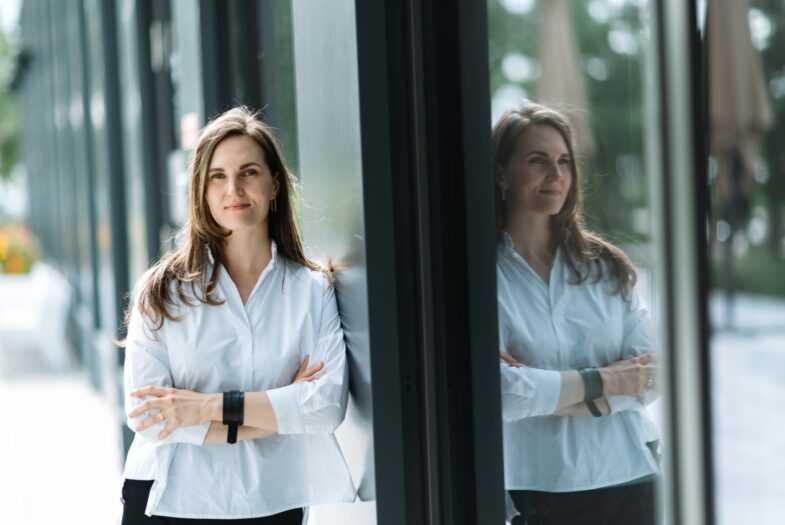 Sieviete baltā kreklā stāv pie mūsdienīgas stikla ēkas sienas ar sakrustotām rokām; stiklā redzams viņas atspulgs, fonā – apstādījumi un gājēju zona.